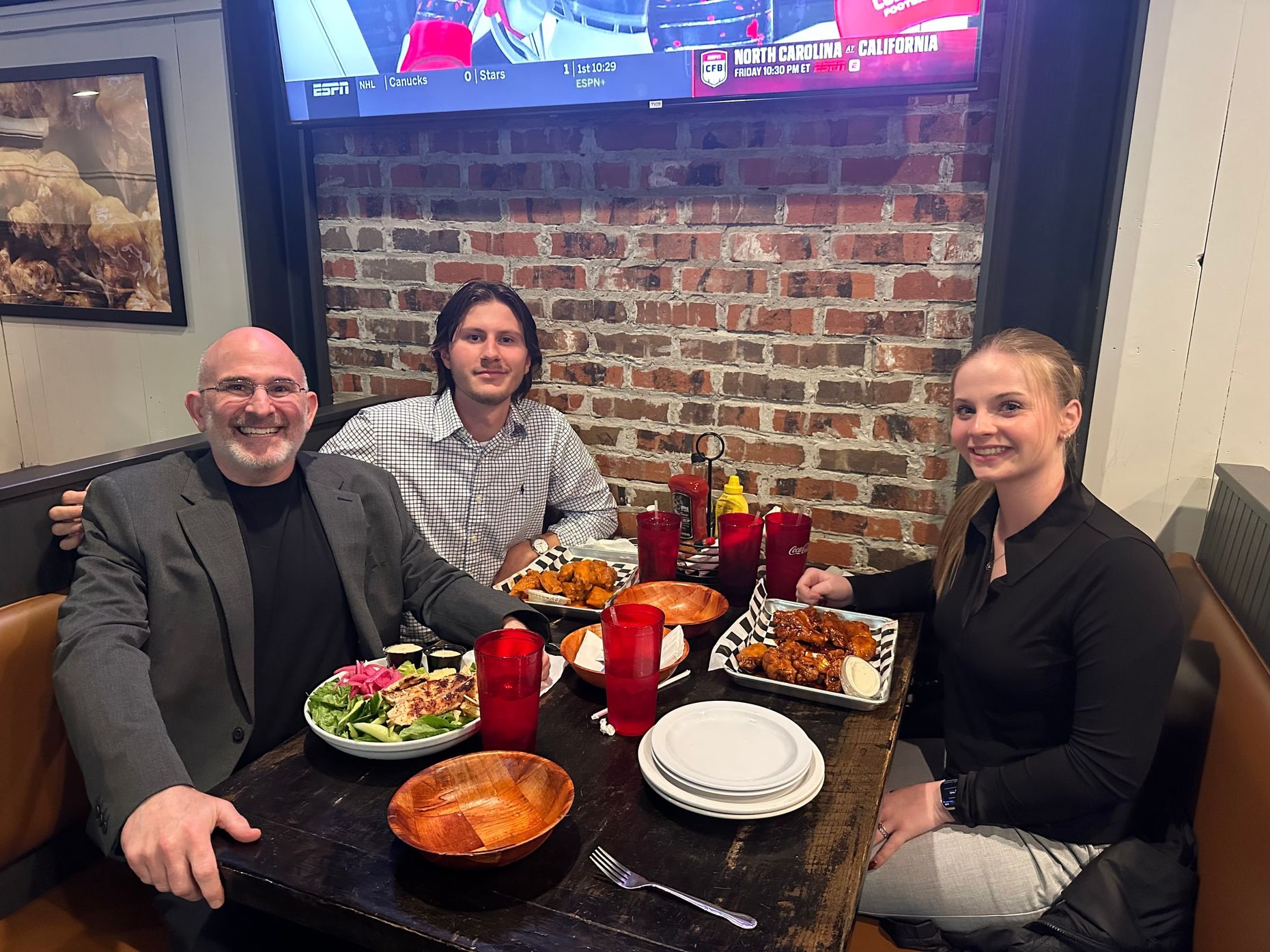 Three people smiling around a restaurant table of wings and salads at BroDop's very first team dinner, October 2025, with the company's first two hires, Kelly Baker and Ryan Roesch.