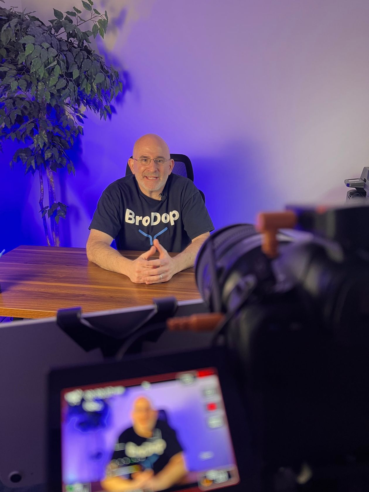 Mark Wolfson at his desk in a BroDop T-shirt, filming a video, seen past the camera with his own image on its screen.