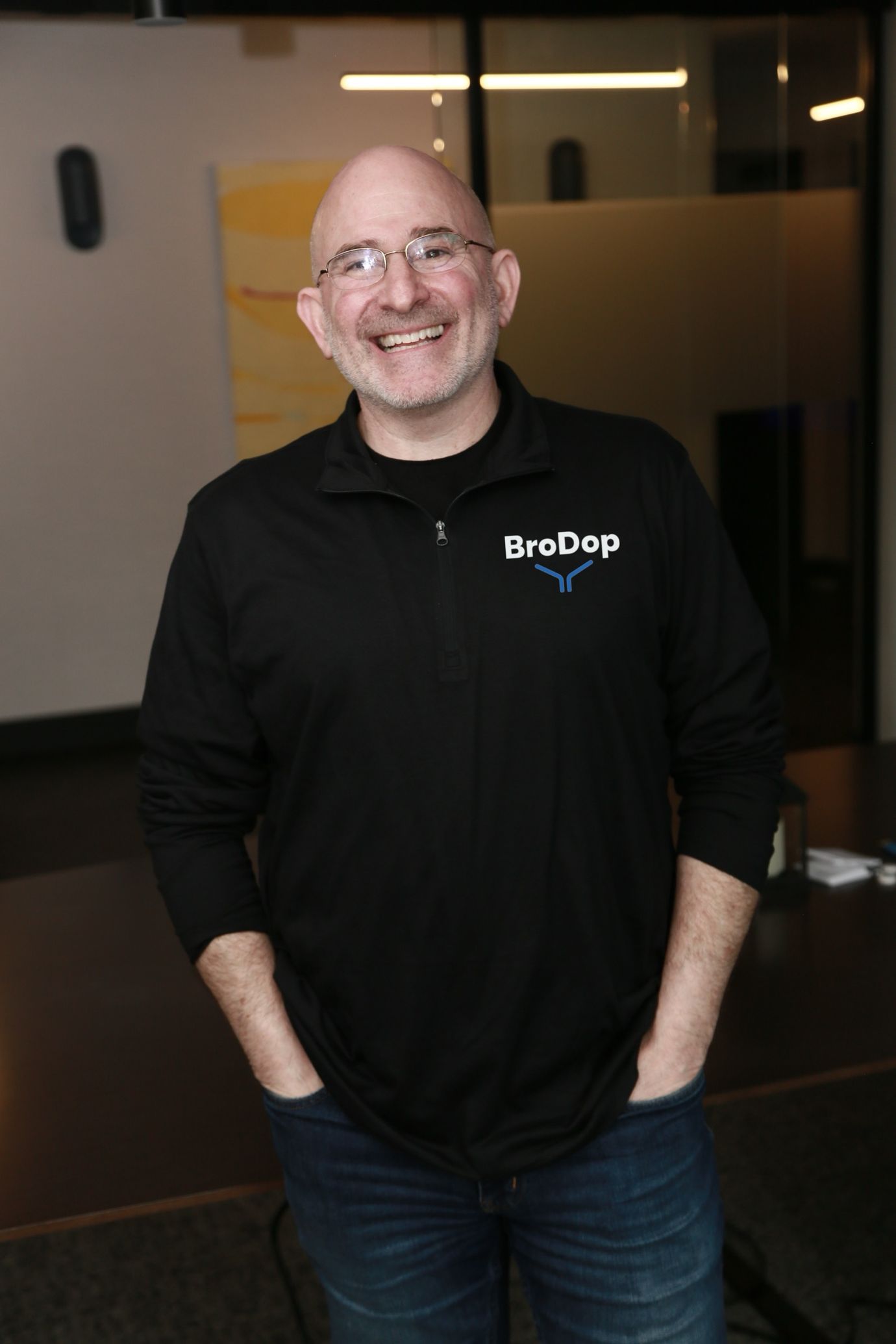Mark Wolfson, smiling with his hands in his pockets, in a black BroDop quarter-zip in a glass-walled office.